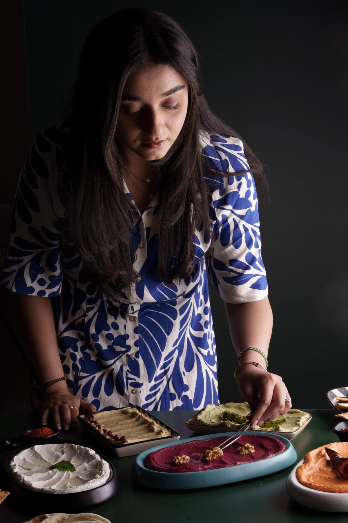 Tanvi in a blue floral dress arranging Saucery dip platters including beetroot hummus and green hummus on a dark background