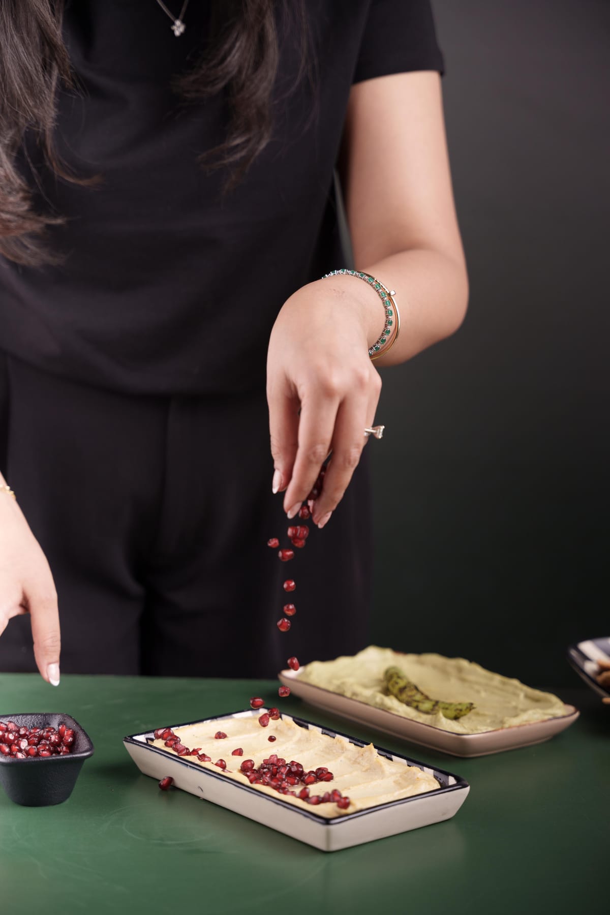 Tanvi sprinkling pomegranate seeds onto a tray of classic hummus, with green hummus tray in background on a dark green surface